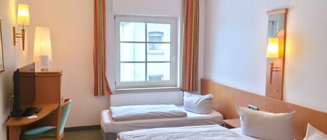 A hotel room with two beds, a desk with a computer, a chair, a trash bin, and a window with curtains.
