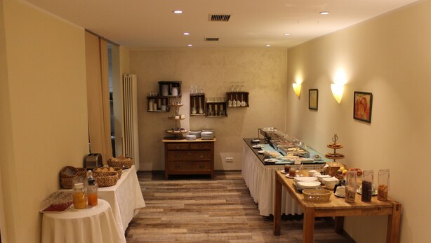 A hotel room with a wooden floor, a dining table with various food items, and a buffet setup with shelves of dishes and condiments. - Hotel Bitburger Hof (Bitburg)
