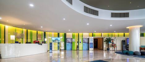 A modern hotel lobby with a curved reception desk, seating area, and a large potted plant.