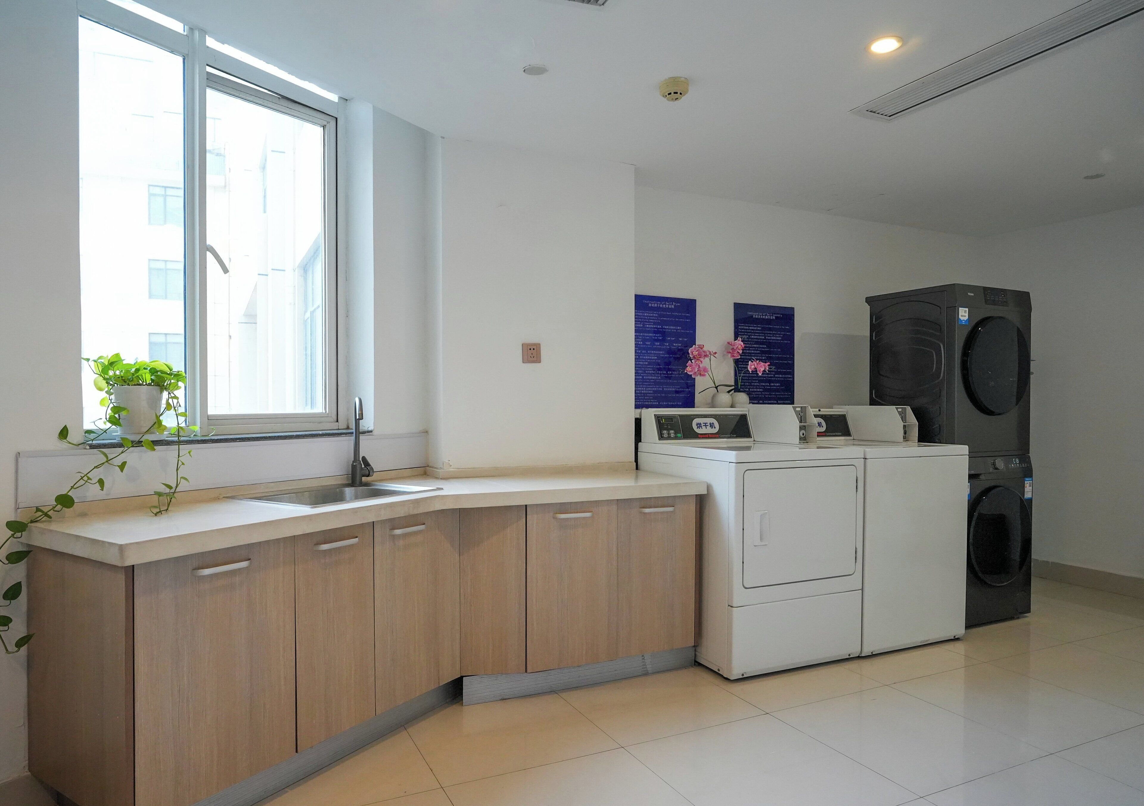 A modern kitchen with a built-in sink, wooden cabinets, a white countertop, a window with a plant, and a stacked washer and dryer.