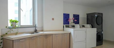 A modern kitchen with a built-in sink, wooden cabinets, a white countertop, a window with a plant, and a stacked washer and dryer.