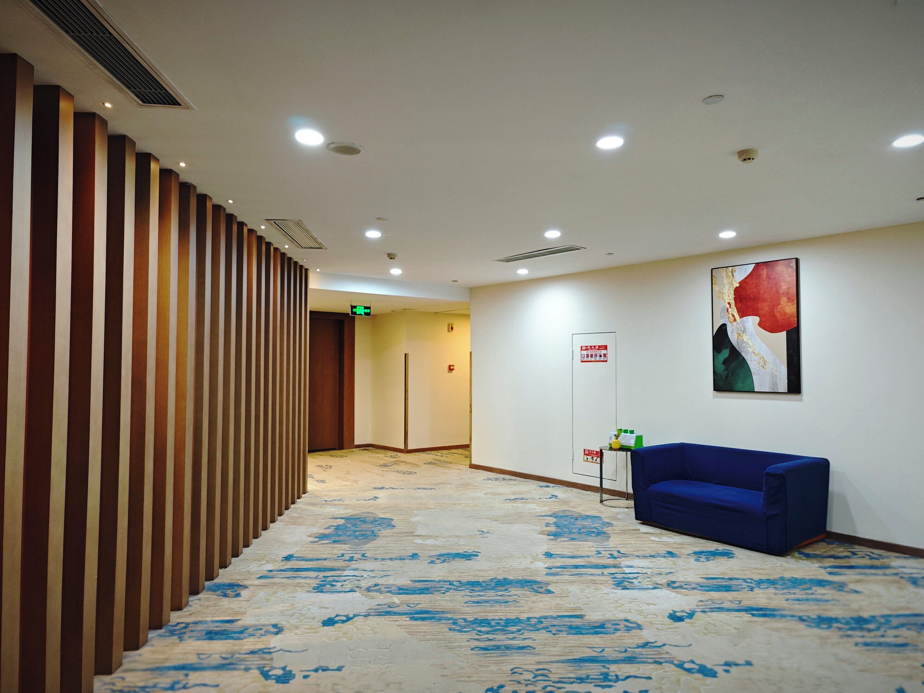 Un hall d’hôtel avec un tapis à motifs, un canapé bleu, une œuvre d’art accrochée au mur et un panneau d’information sur la sécurité incendie.
