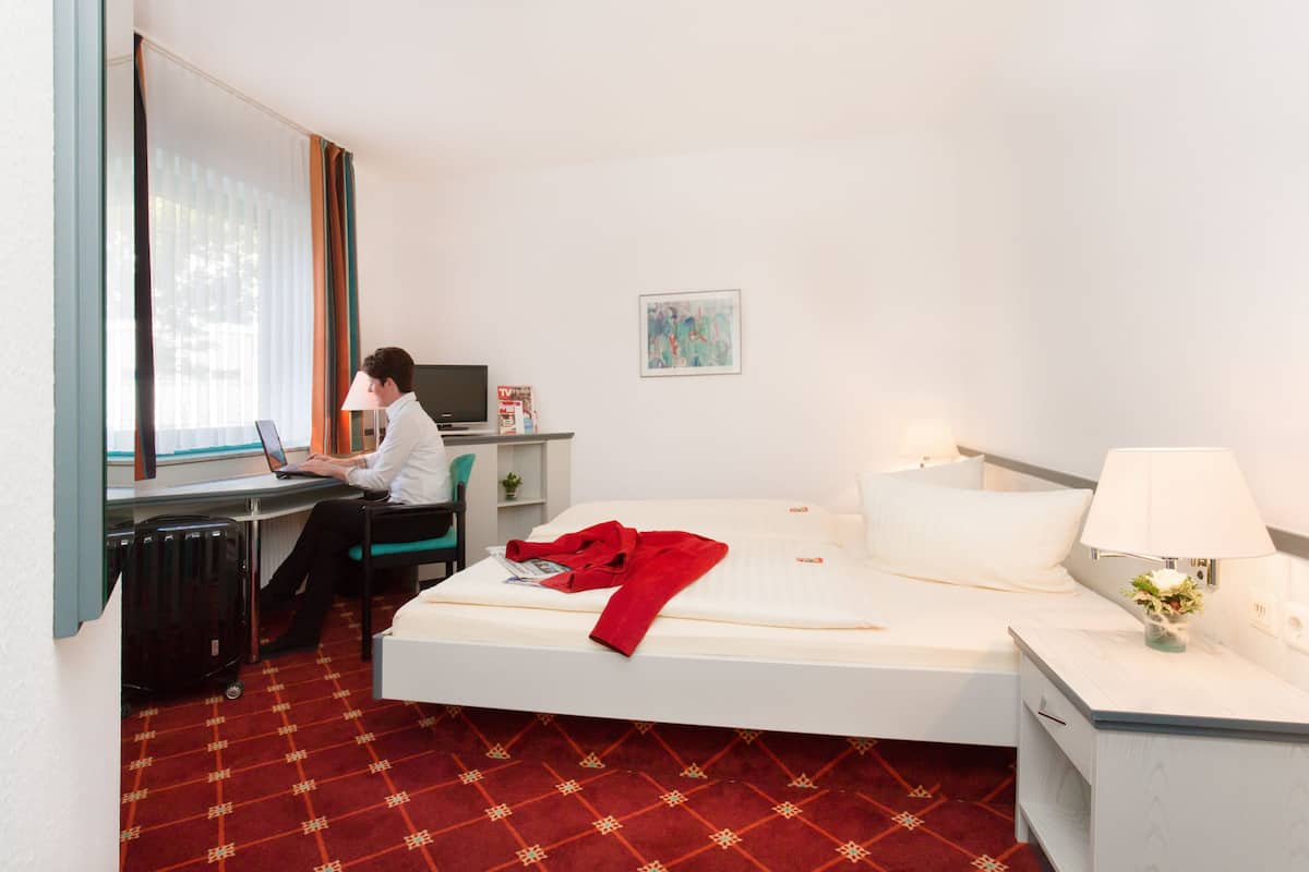 Une chambre d’hôtel avec un lit, un bureau avec un ordinateur portable, une chaise, une valise, une lampe et un tableau au mur.