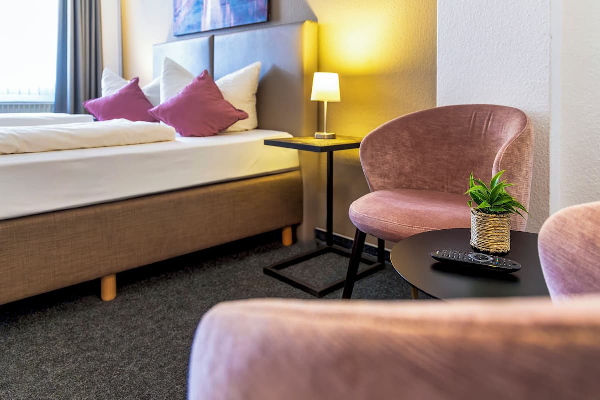 A hotel room with a bed, a small table with a lamp, a chair, and a potted plant.