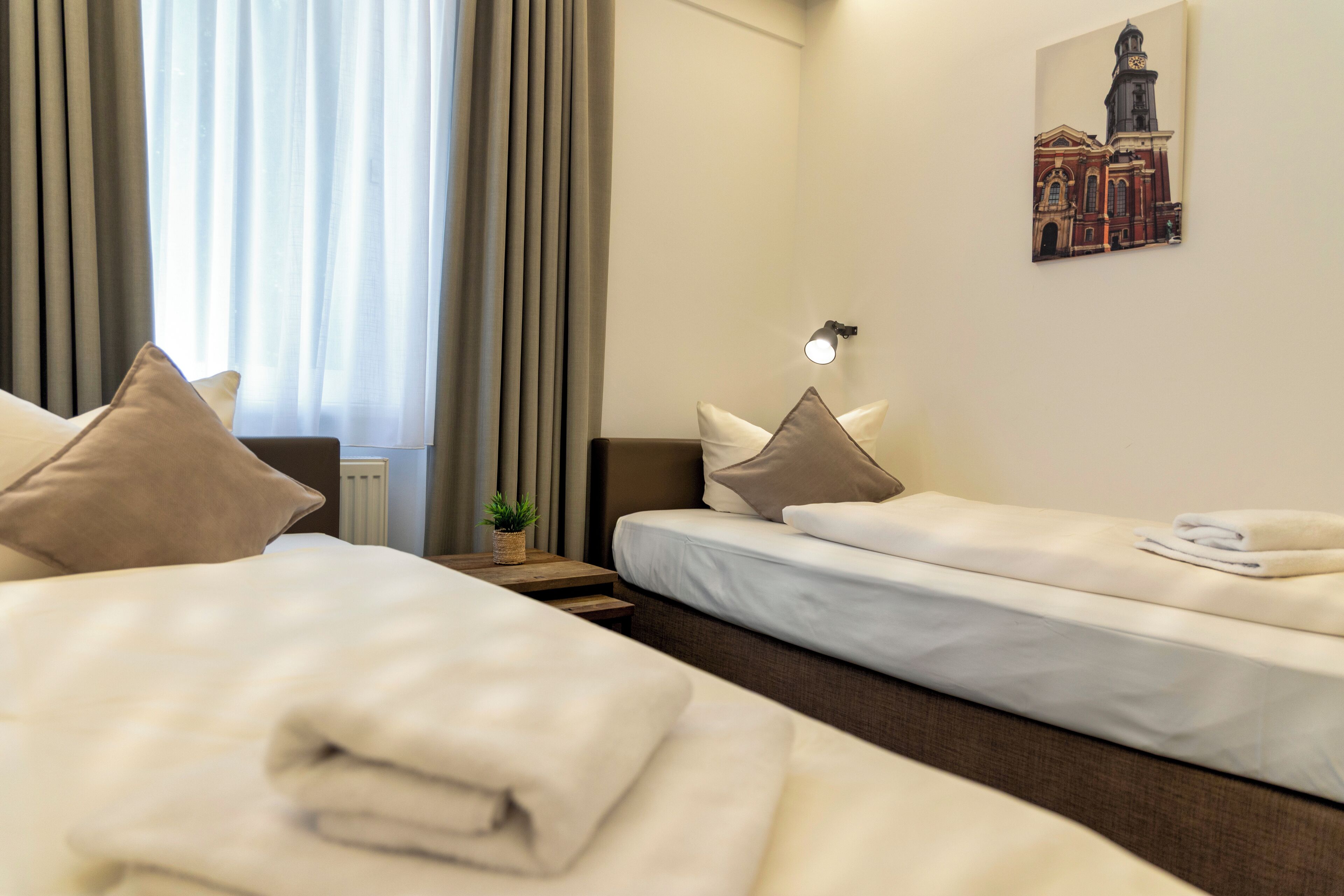 A hotel room with two beds, a nightstand with a lamp, a window with curtains, and a painting on the wall.