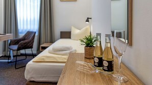 A hotel room with a bed, a desk, a chair, a bedside table with wine bottles and glasses, and a plant. - Auto-Parkhotel (Hamburg)