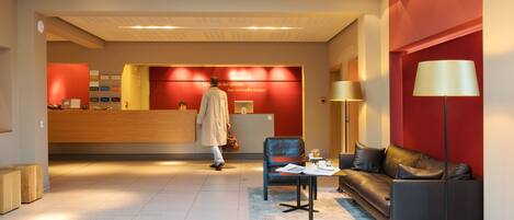 A modern hotel lobby with a reception desk, seating area, and a large window.