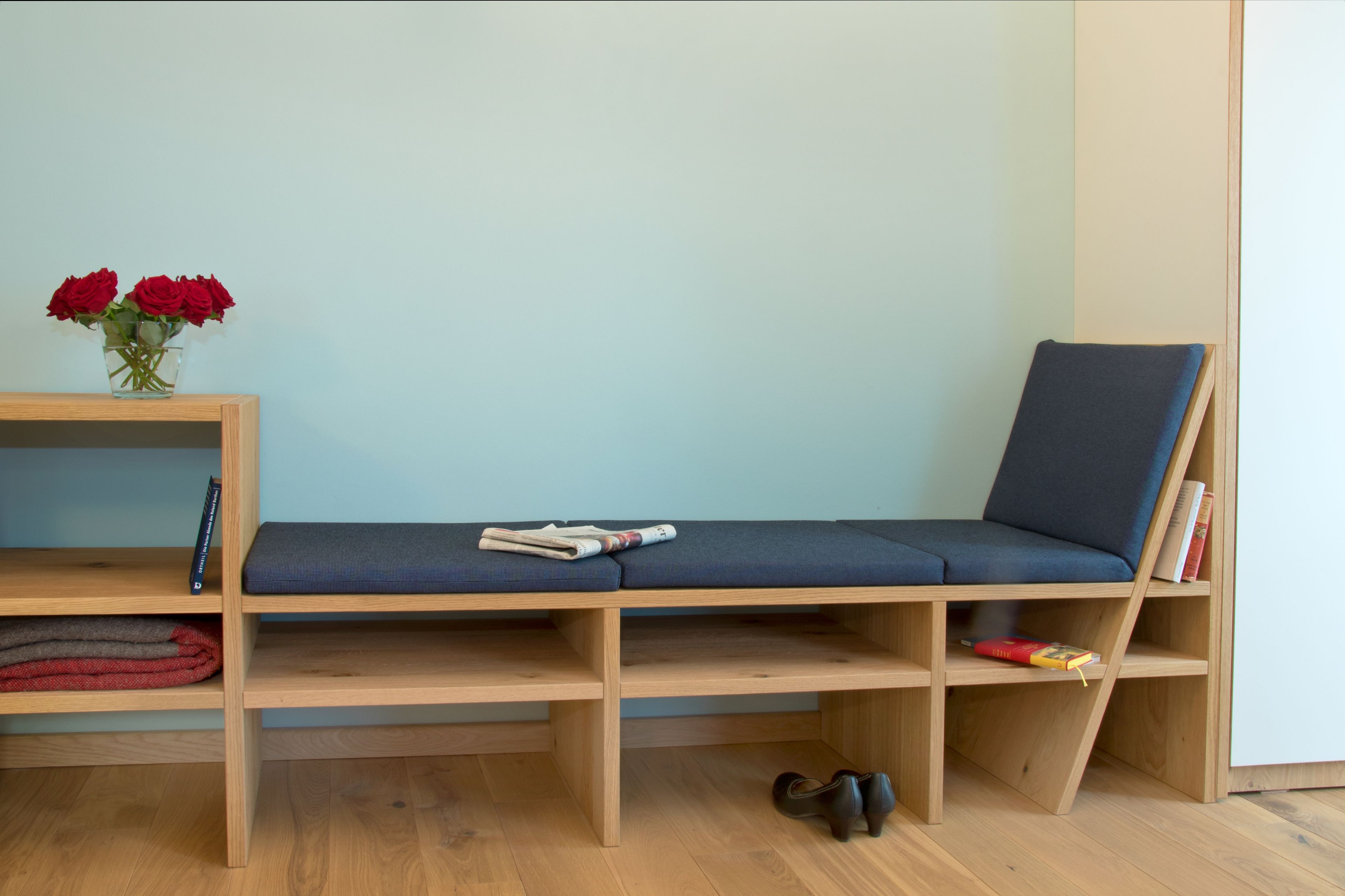 Een kamer met een houten bank met een blauw kussen, een houten plank met boeken en een vaas rode rozen, en een paar schoenen op de vloer.