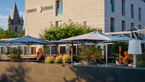 Un hôtel à la façade moderne, doté d’une terrasse avec des parasols, et dont l’arrière-plan est dominé par une structure évoquant un château.