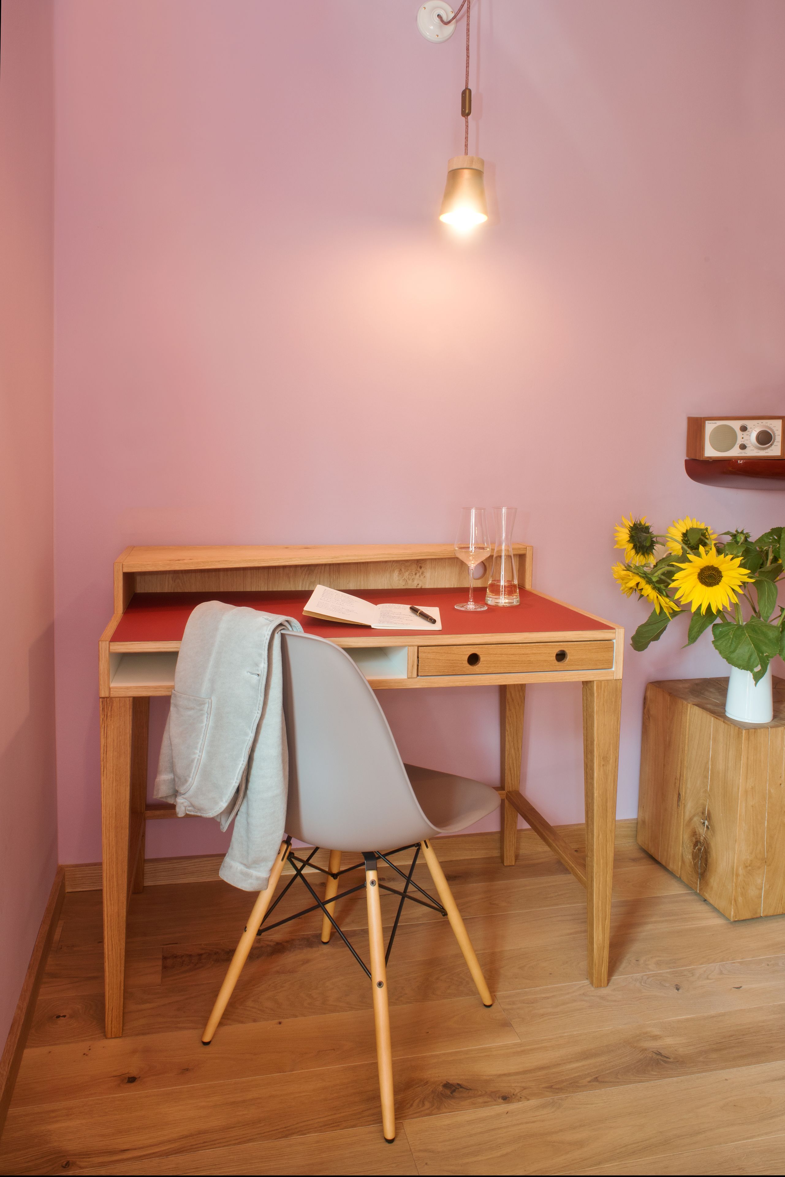 Un bureau en bois avec un plateau rouge, une chaise blanche, une lampe suspendue, une radio et un vase rempli de tournesols.