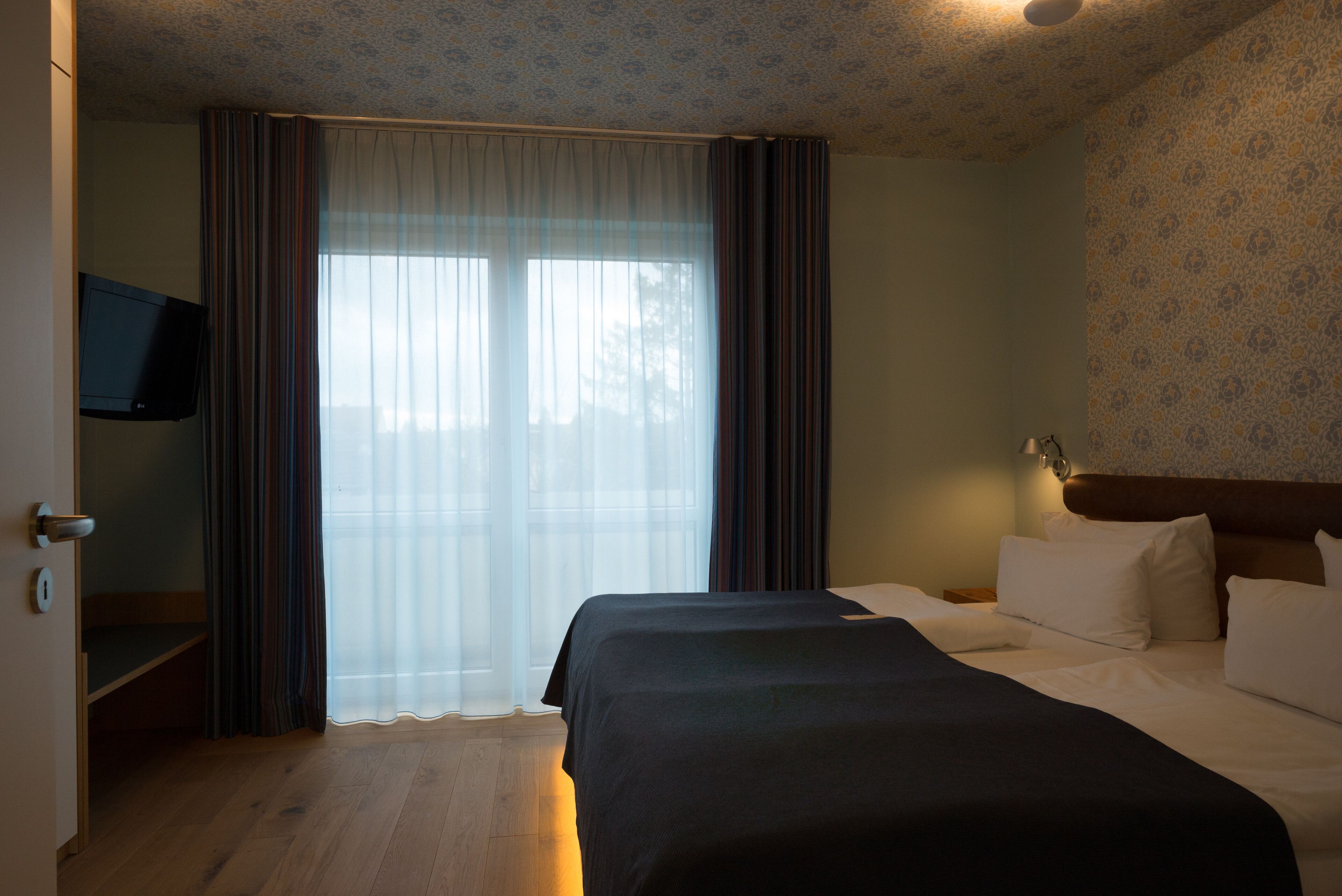 A hotel room with a bed, a television mounted on the wall, and a window with curtains.