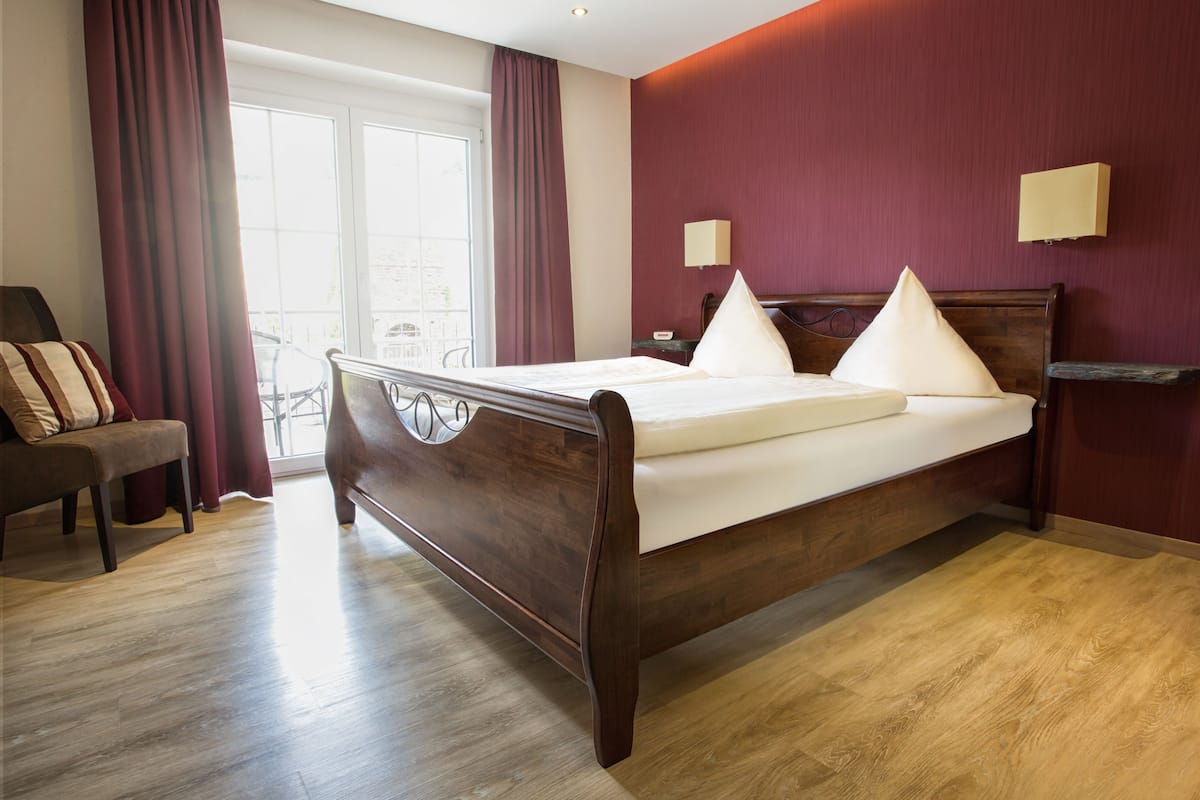 A hotel room with a wooden bed, a chair, a window with curtains, and a wall-mounted lamp.