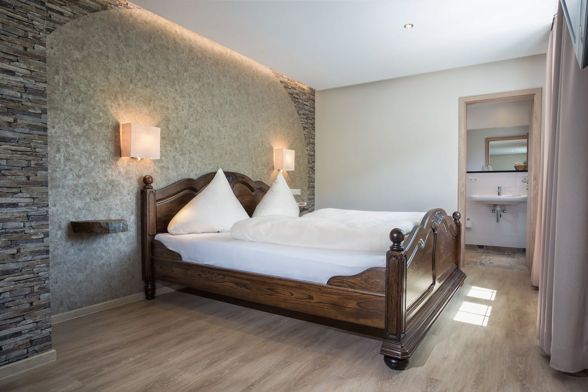 A bedroom with a wooden bed, stone wall, and a bathroom visible through an open door.