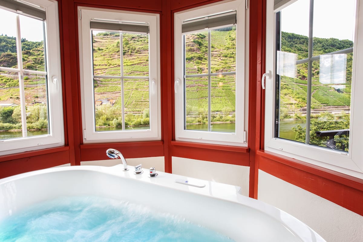 A bathroom with a large bathtub, three windows, and a view of a vineyard.