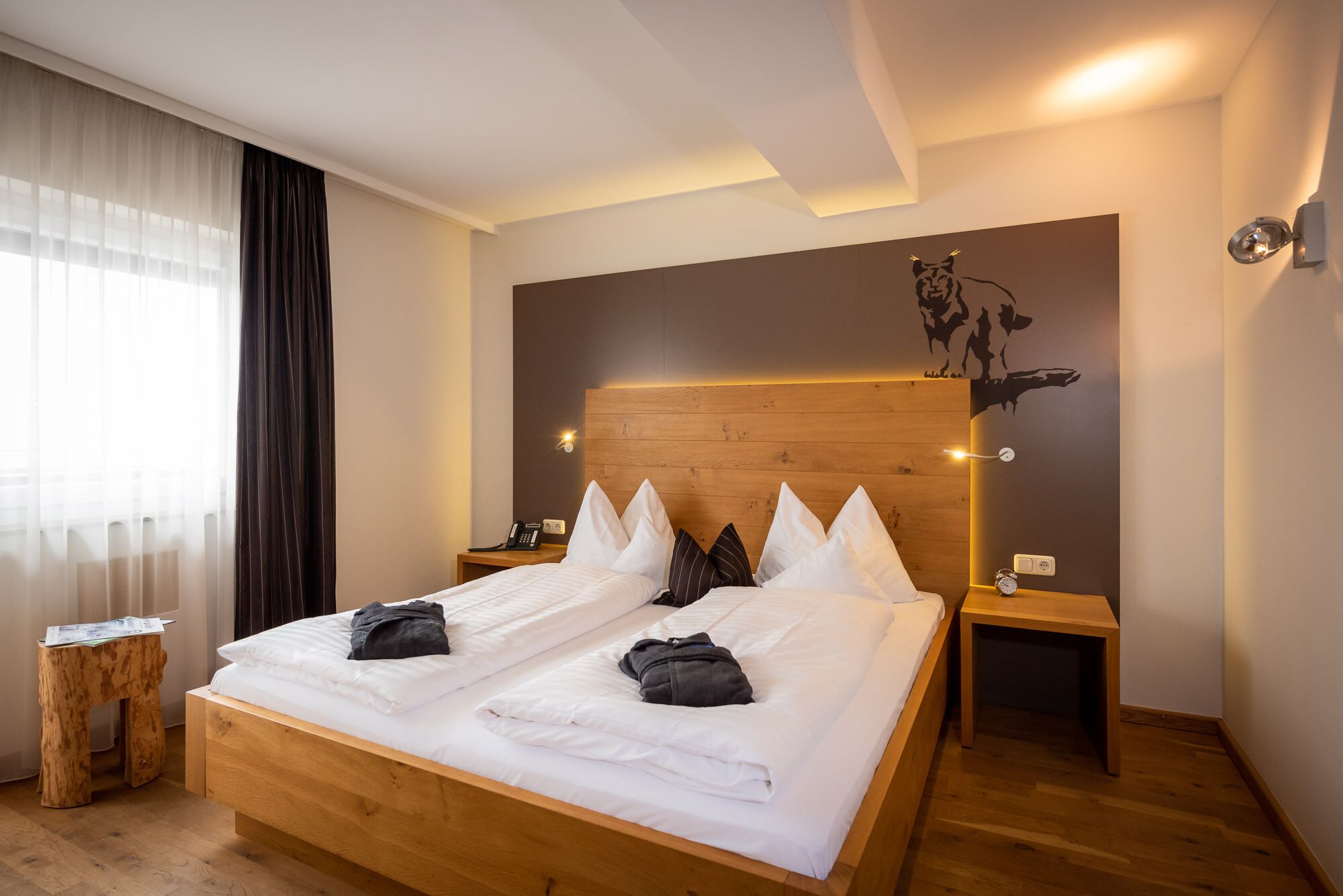 A hotel room with a wooden bed, white bedding, a wooden headboard, a wall-mounted artwork, a bedside table with a lamp, and a small wooden stool.
