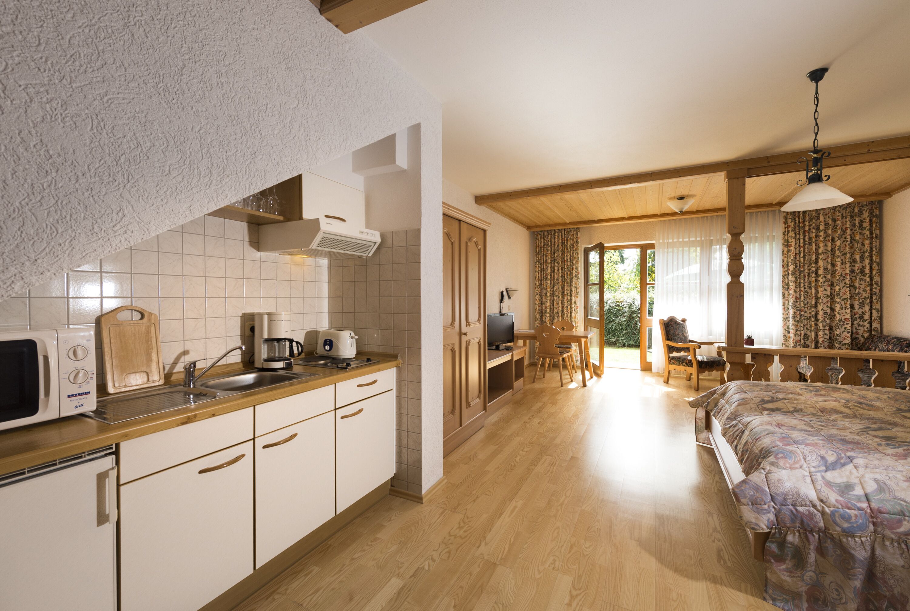 A compact kitchen with a microwave, toaster, and coffee maker, a dining area with a table and chairs, and a bedroom with a bed and curtains.