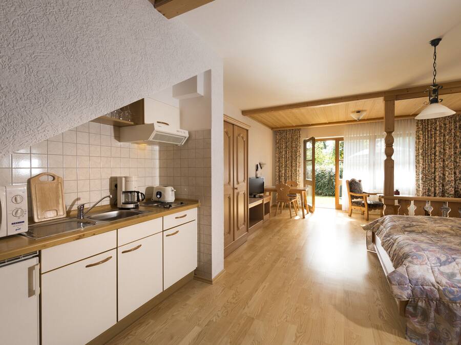 A compact kitchen with a microwave, toaster, and coffee maker, a dining area with a table and chairs, and a bedroom with a bed and curtains.