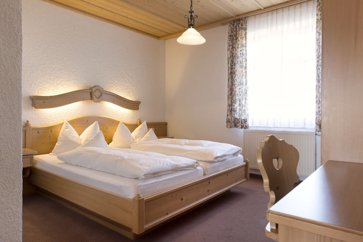 A double bed with a wooden headboard, a bedside table, and a chair in a room with a window and a radiator.