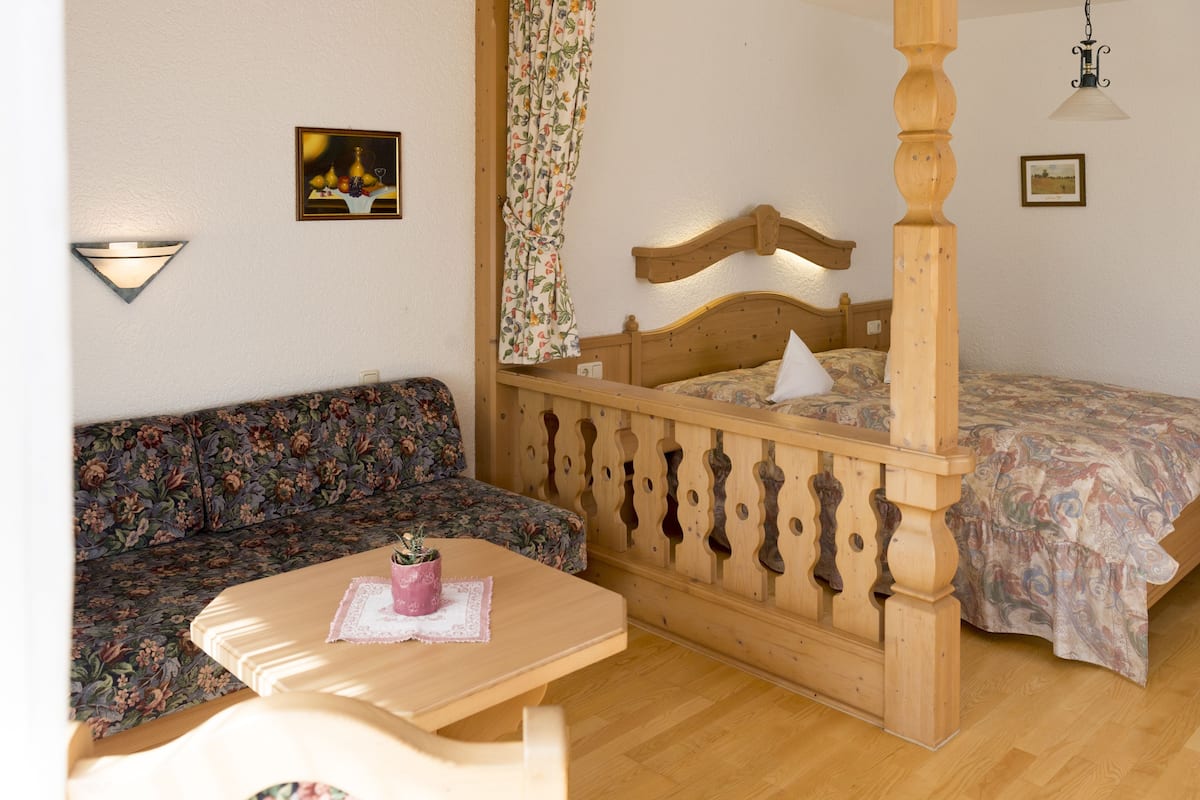 A bedroom with a four-poster bed, a floral sofa, a wooden table with a pink candle, and a wall-mounted lamp.