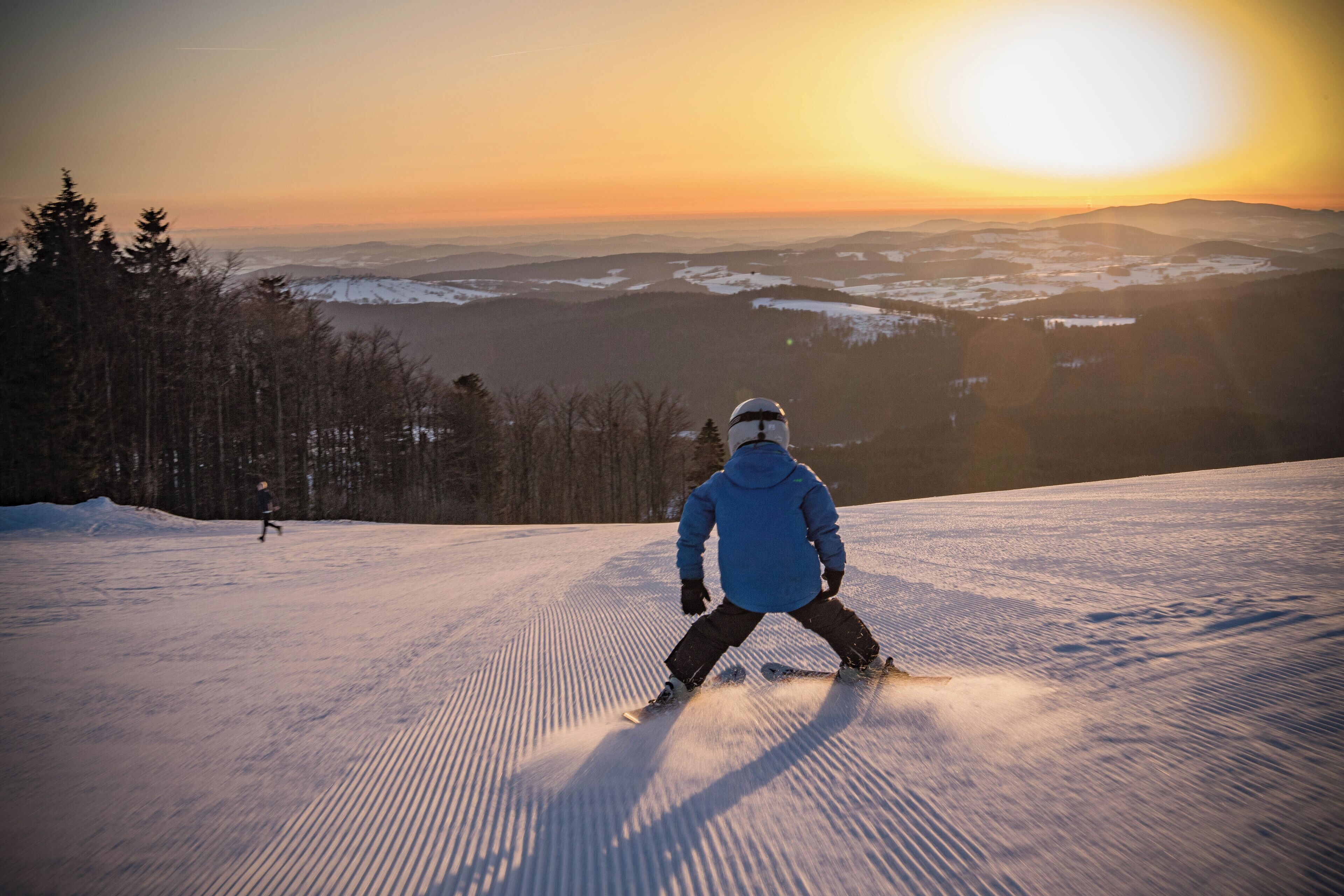 Een persoon skiënd een sneeuwachtig helling met een prachtig uitzicht op bergen en een ondergaande zon op de achtergrond.