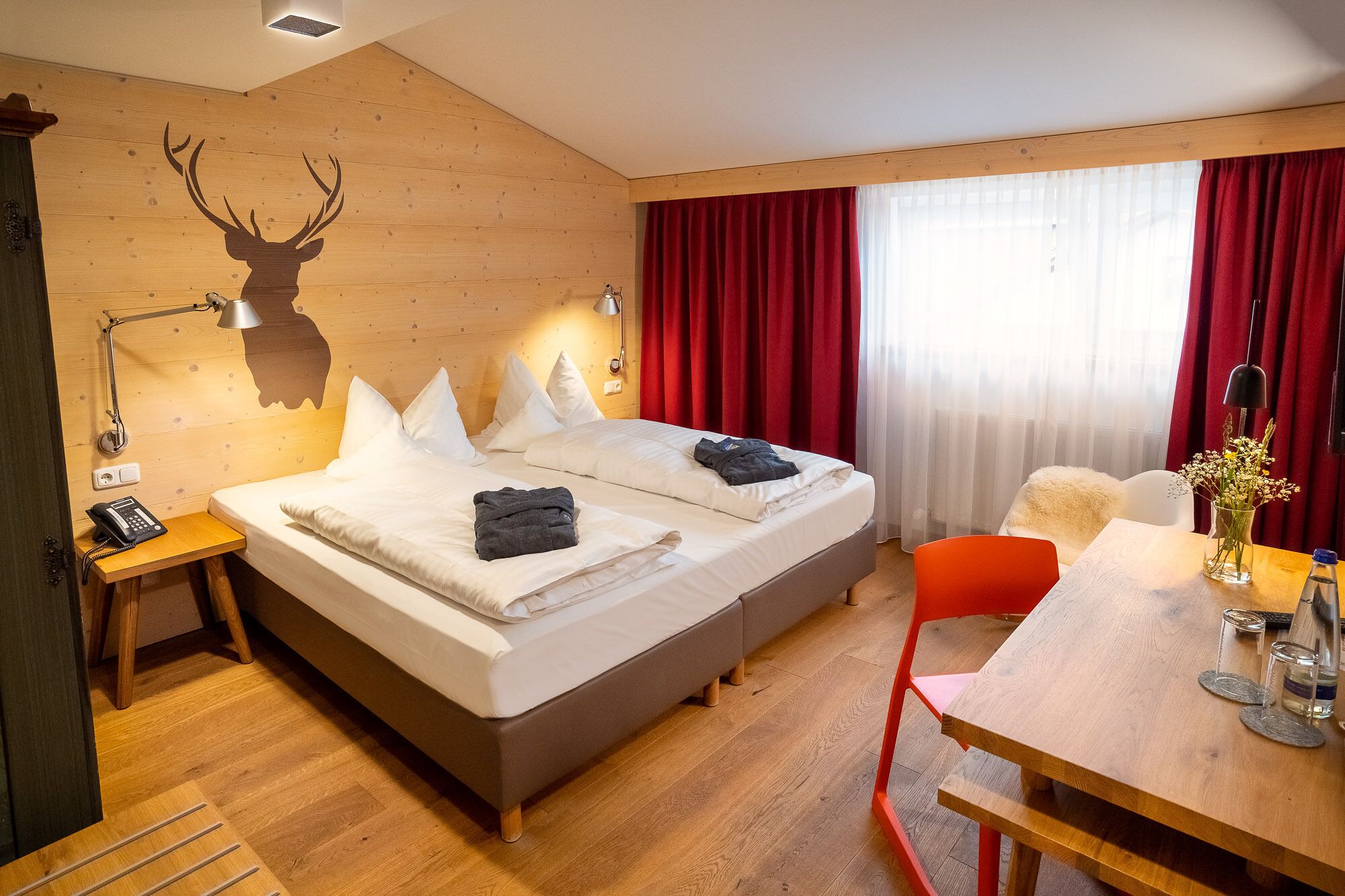 A hotel room with a wooden wall, a bed with white bedding, a red curtain, a table with a vase of flowers, and a chair.