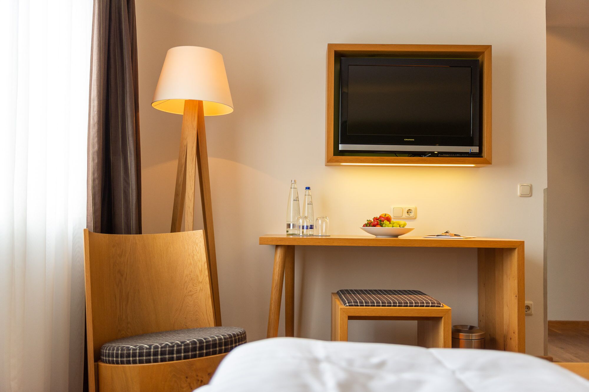 Ein Hotelzimmer mit einem Holztisch, einem Fernseher, einer Lampe, einer Obstschale und einem Sessel mit kariertem Kissen.