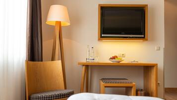 Ein Hotelzimmer mit einem Holztisch, einem Fernseher, einer Lampe, einer Obstschale und einem Sessel mit kariertem Kissen.
