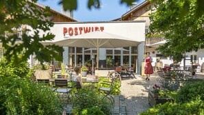 An outdoor seating area with tables and chairs, a large umbrella, and a building with a sign that reads "POSTWIRT". - Landhotel Postwirt (Grafenau)