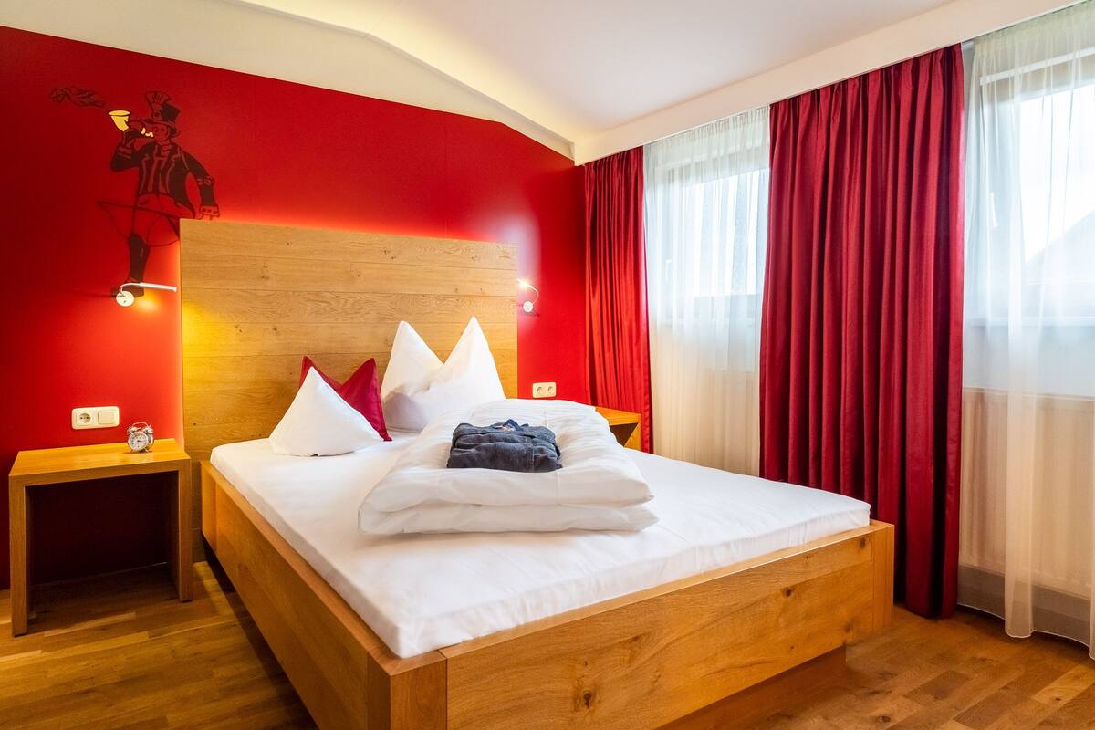 A neatly made bed with white linens and a blue towel, a wooden headboard, a red accent wall with a mural, and a window with red curtains.