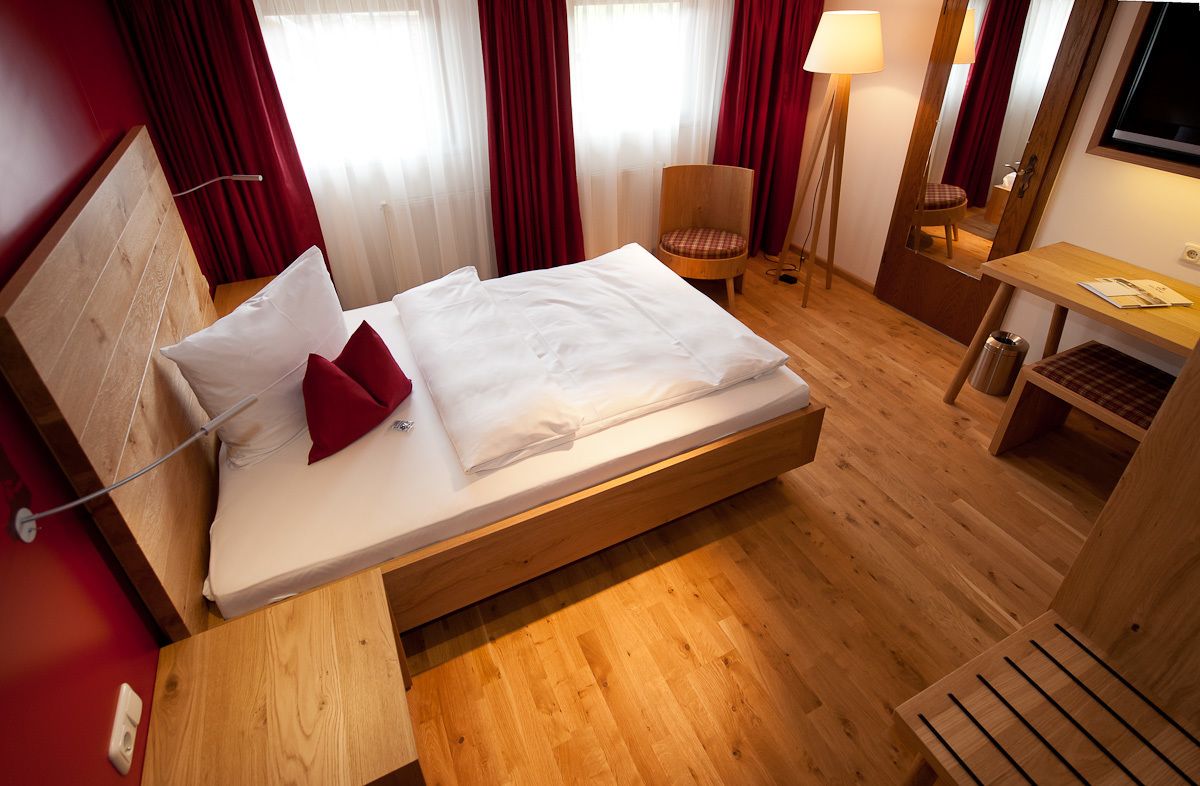 A hotel room with a wooden bed, red curtains, a chair, a desk, and a TV.