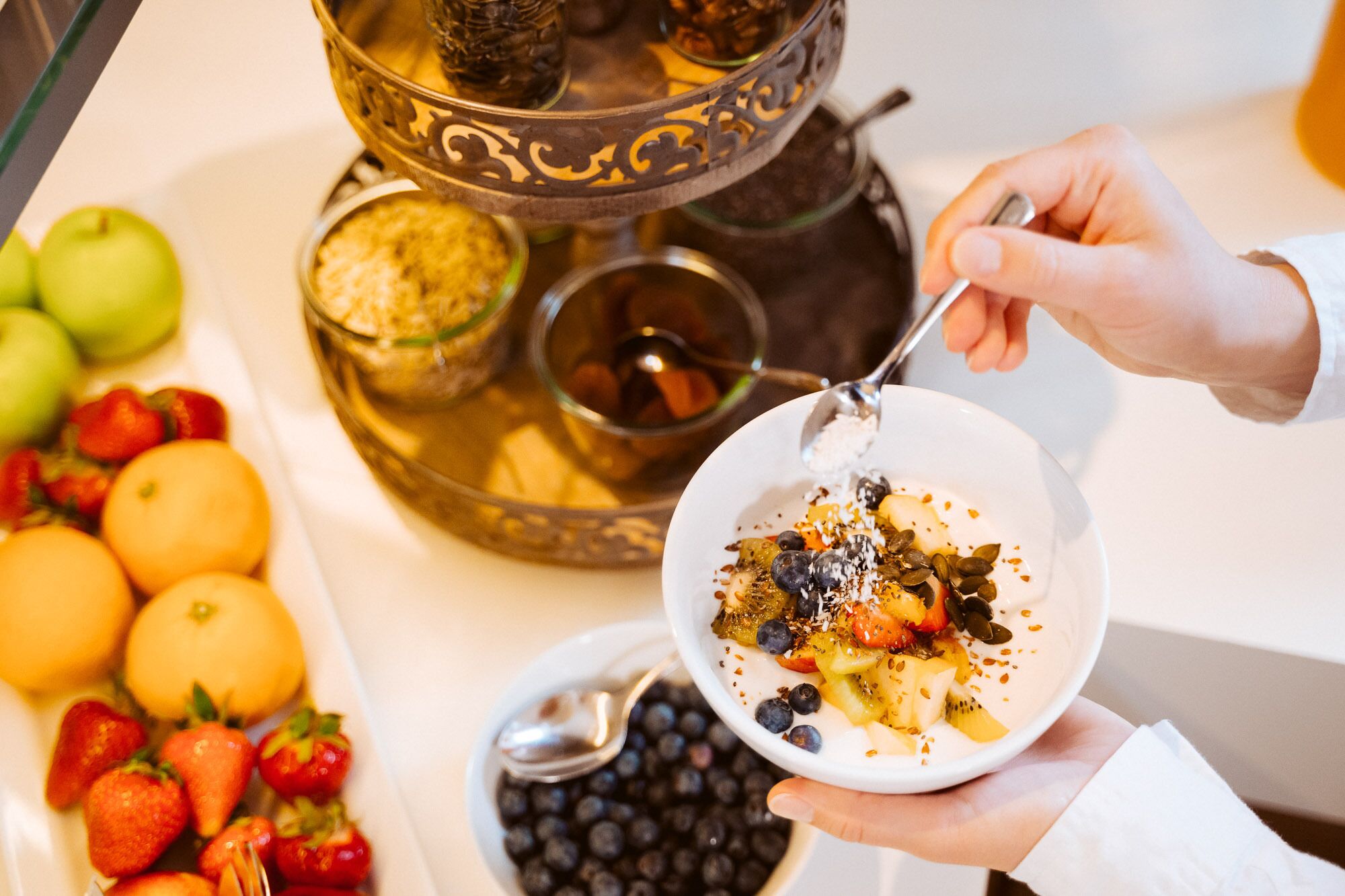A person holding a bowl of yogurt with fruit and seeds, with a tray of fruits and a decorative metal stand in the background.