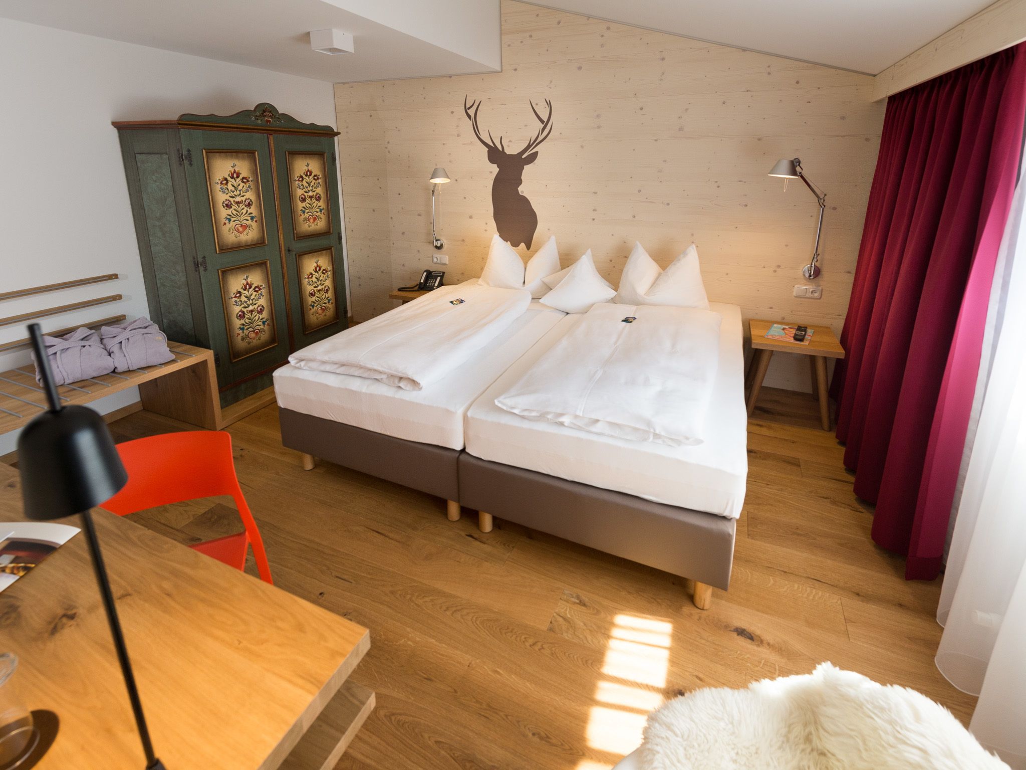 Ein Doppelbett mit weißer Bettwäsche, ein Holzkopfschild, ein Nachttisch mit floralen Verzierungen, ein Schreibtisch mit Stuhl und ein roter Vorhang.