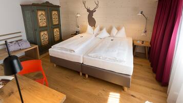 Ein Doppelbett mit weißer Bettwäsche, ein Holzkopfschild, ein Nachttisch mit floralen Verzierungen, ein Schreibtisch mit Stuhl und ein roter Vorhang.