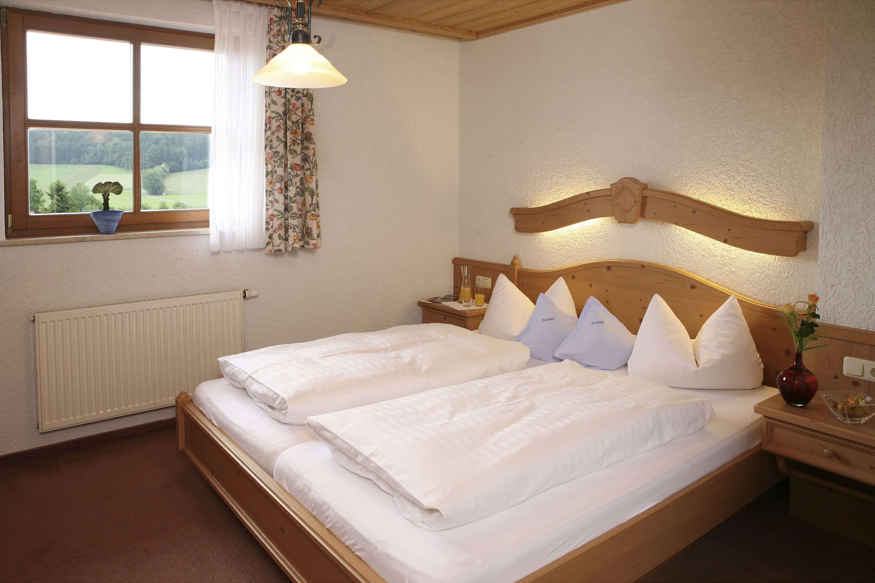 Un lit double avec du linge de lit blanc, une tête de lit en bois, une table de chevet avec une lampe, et une vue sur un paysage verdoyant par la fenêtre.