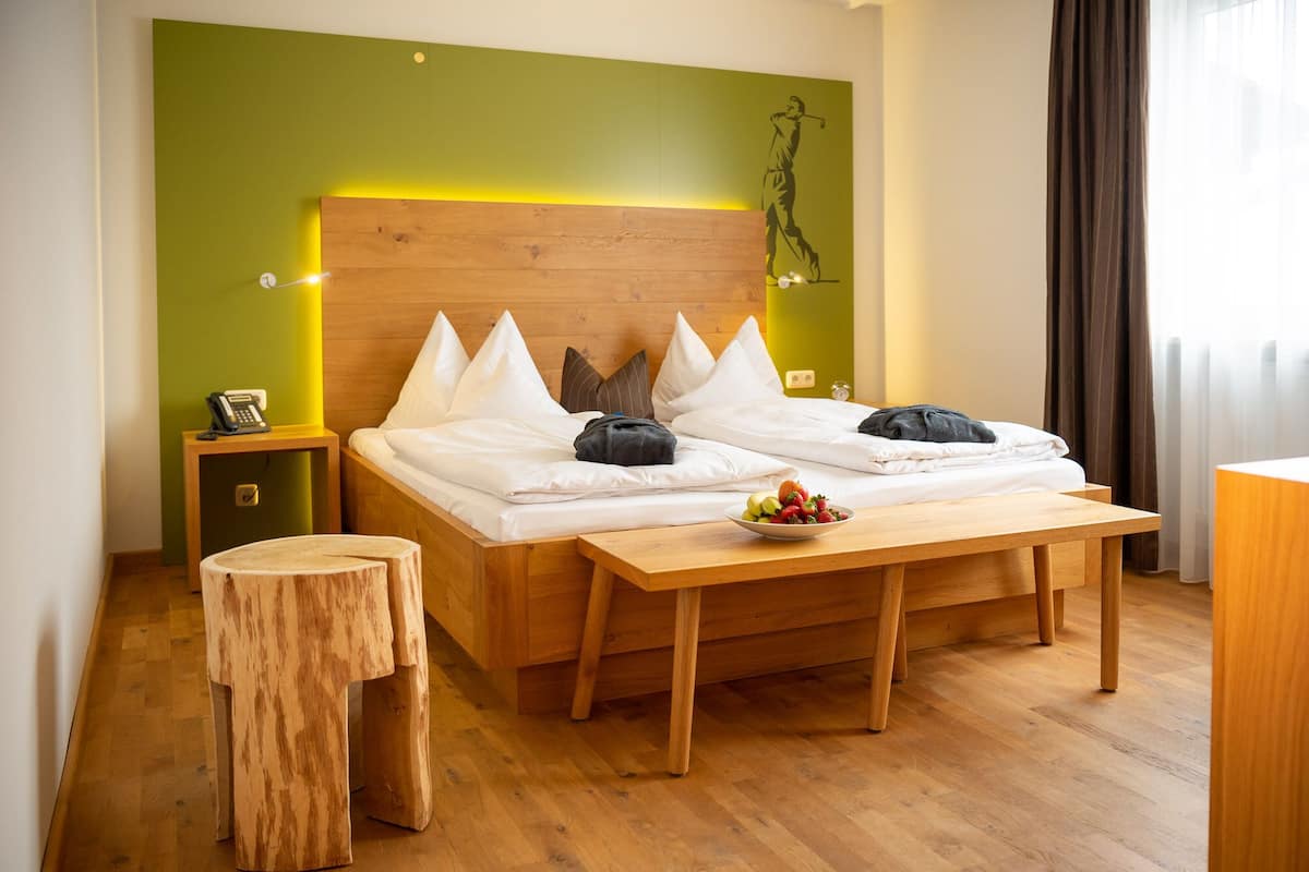A hotel room with a wooden bed, a bedside table with a phone, a small wooden stool, and a fruit bowl on the bed.