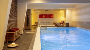 A modern indoor swimming pool with a wooden bench and a candle on the floor, a chair, and a red abstract painting on the wall. - Landhotel Postwirt (Grafenau)
