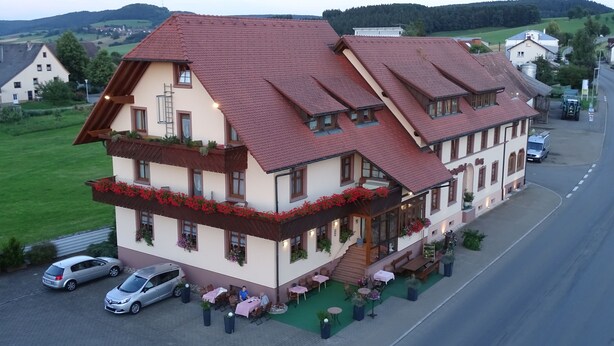 A traditional European-style building with a red-tiled roof, multiple balconies with flower boxes, and a paved outdoor area with tables and chairs.