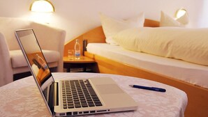 A hotel room with a laptop on a round table, a bed with white linens, a bedside table with a water bottle, and a lamp on the wall.