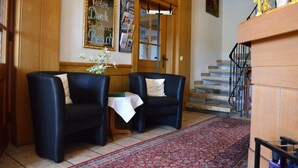 A room with two black armchairs, a small table with a white tablecloth, a red patterned rug, wooden paneling, a staircase, and a framed sign on the wall.