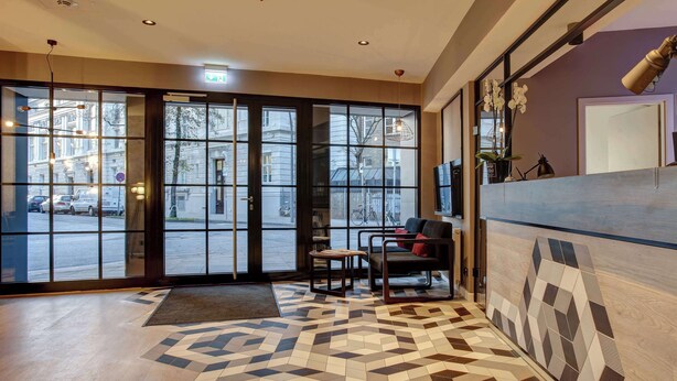 En hotellobby med ett golv i geometriskt mönster, stora glasdörrar som leder ut och en receptionsdisk med modern design.