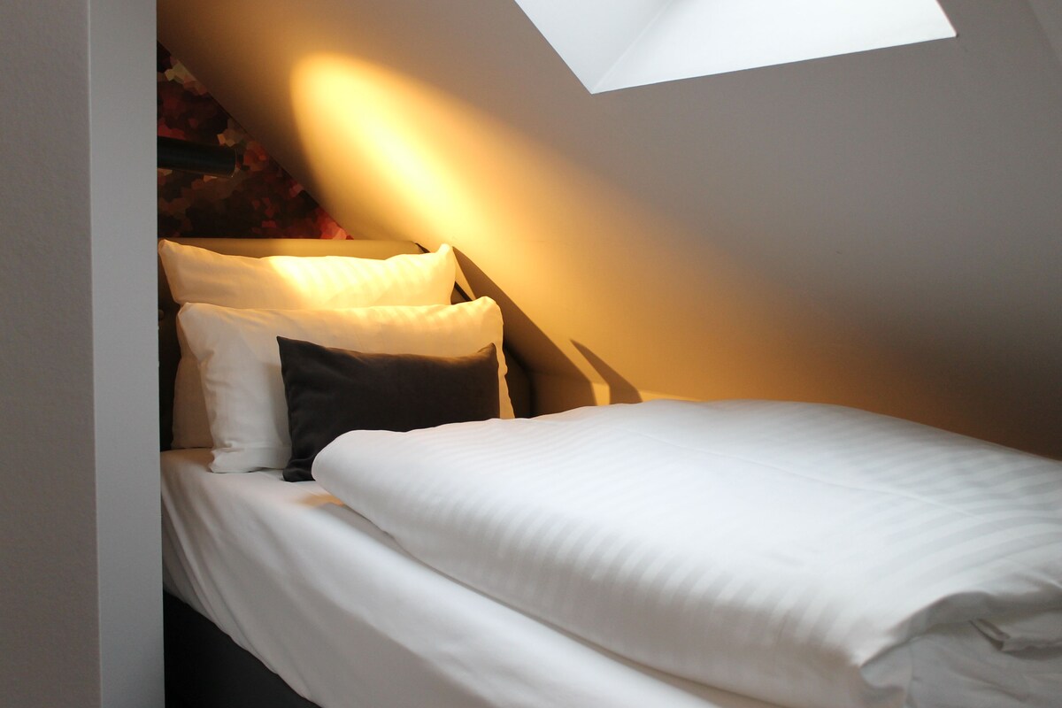 Ein kleines, gemütliches Schlafzimmer mit einem Einzelbett, einem Oberlicht und einem teilweisen Blick auf eine Wand mit einem Muster.