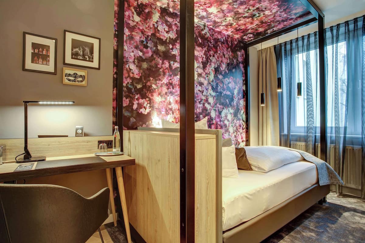 Una habitación de hotel con un escritorio de madera, una cama con ropa blanca y un papel tapiz floral, además de vistas al exterior.
