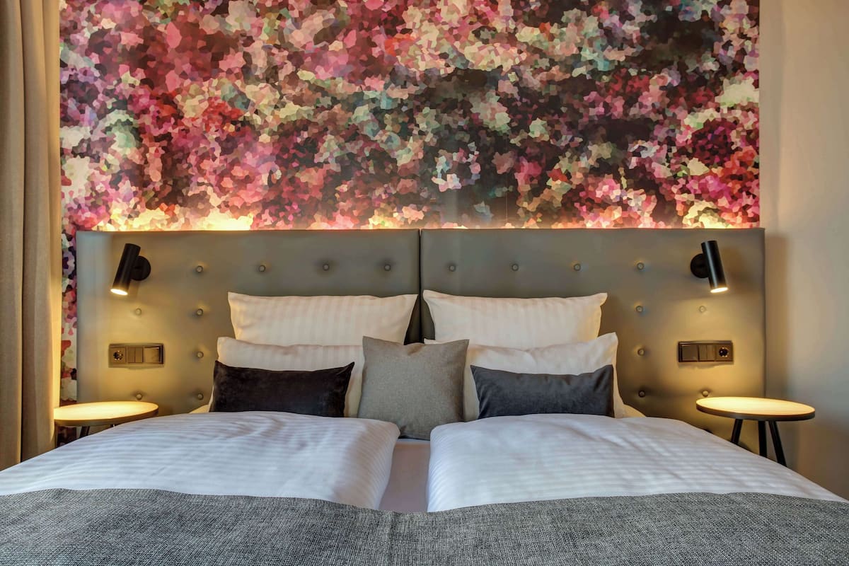 Una habitación de hotel con una cama grande, un cabecero con un cuadro floral, dos mesitas de noche y una pequeña mesa redonda.