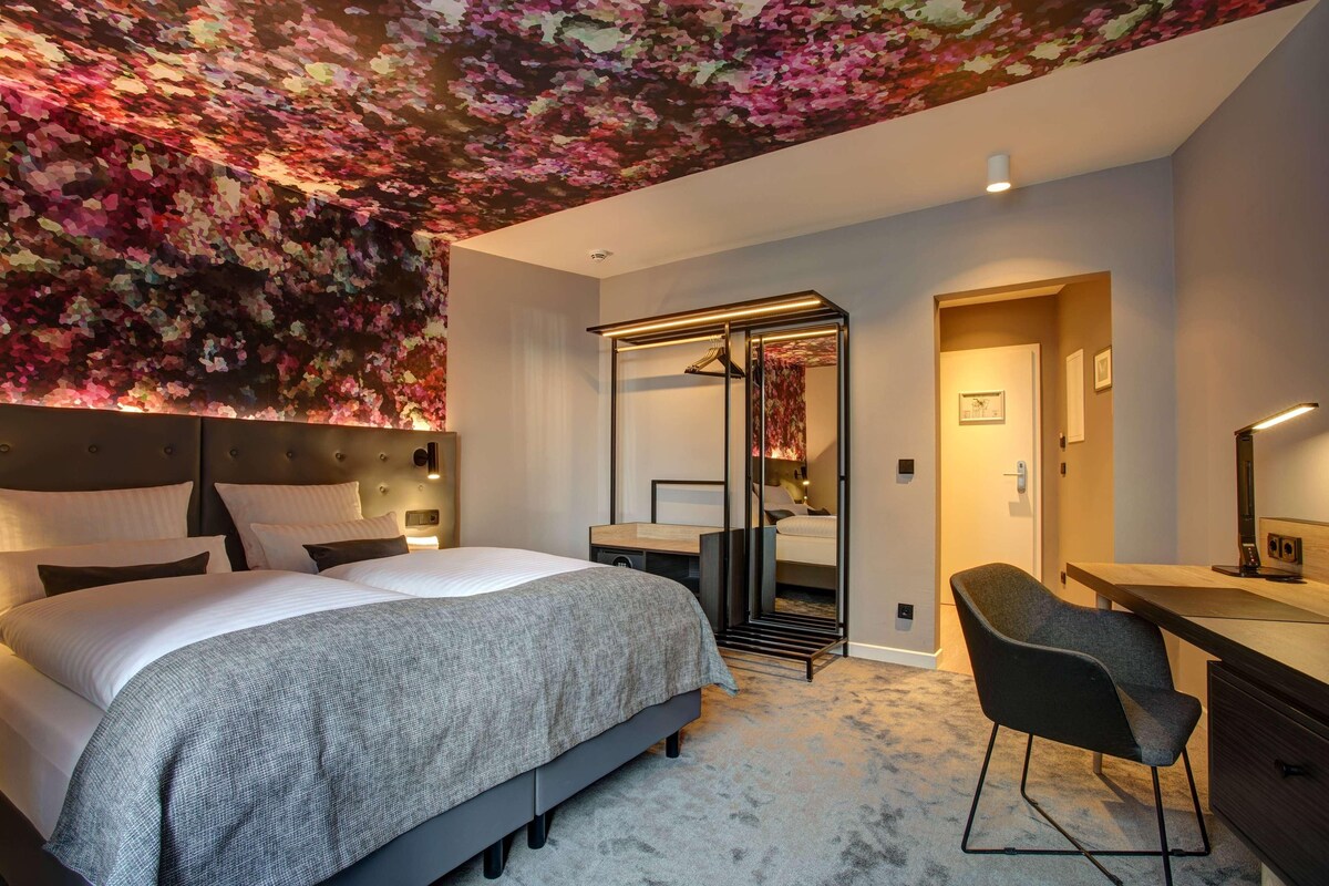 Ein modernes Hotelzimmer mit einem großen Bett, einem Schreibtisch und einem Stuhl. Ein Kleiderschrank mit Spiegel und eine Tür zu einem weiteren Raum sind ebenfalls vorhanden.