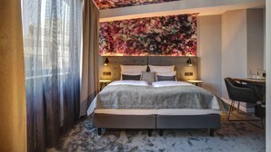 Ett hotellrum med en stor säng, ett skrivbord och en stol. Rummet har ett blommigt mönster i taket och ett fönster med tunna, genomskinliga gardiner.