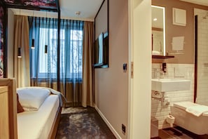 Habitación de hotel con cama, televisor, baño con lavabo e inodoro, y ventana con cortinas.