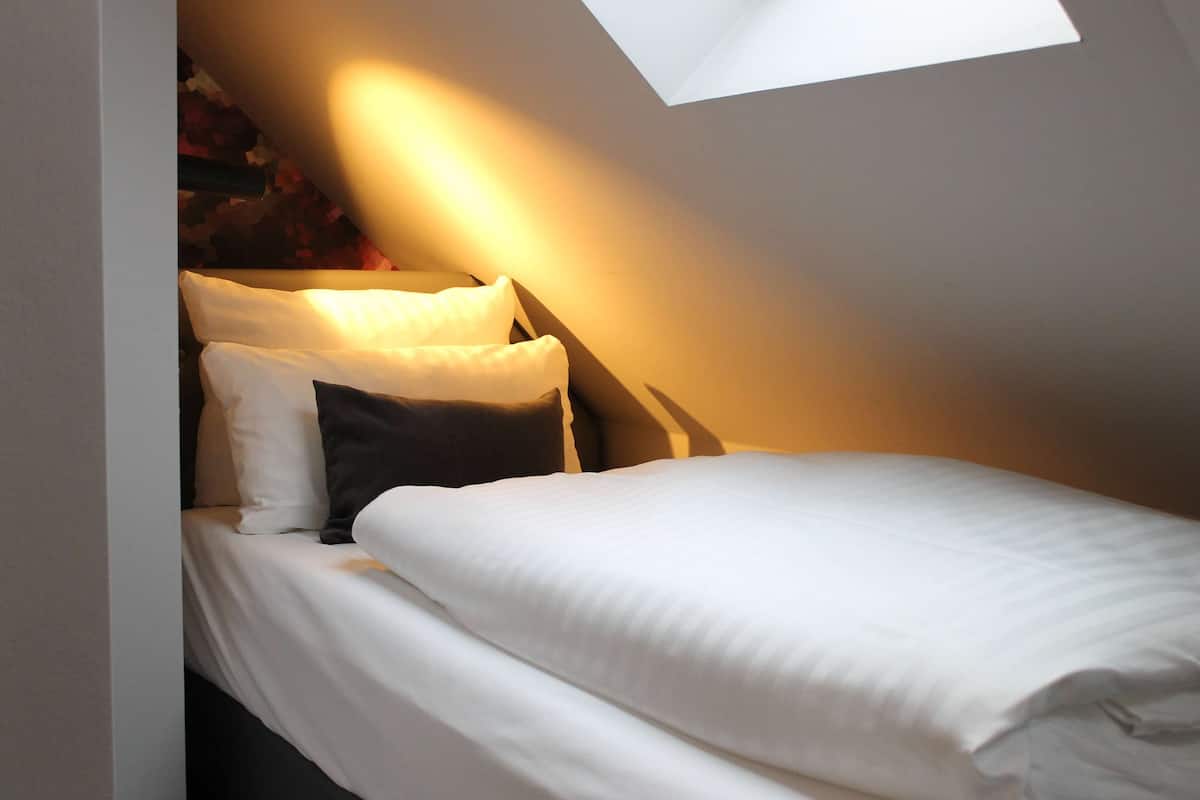 Una cama con ropa de cama blanca y un almohadón, techo inclinado y tragaluz.