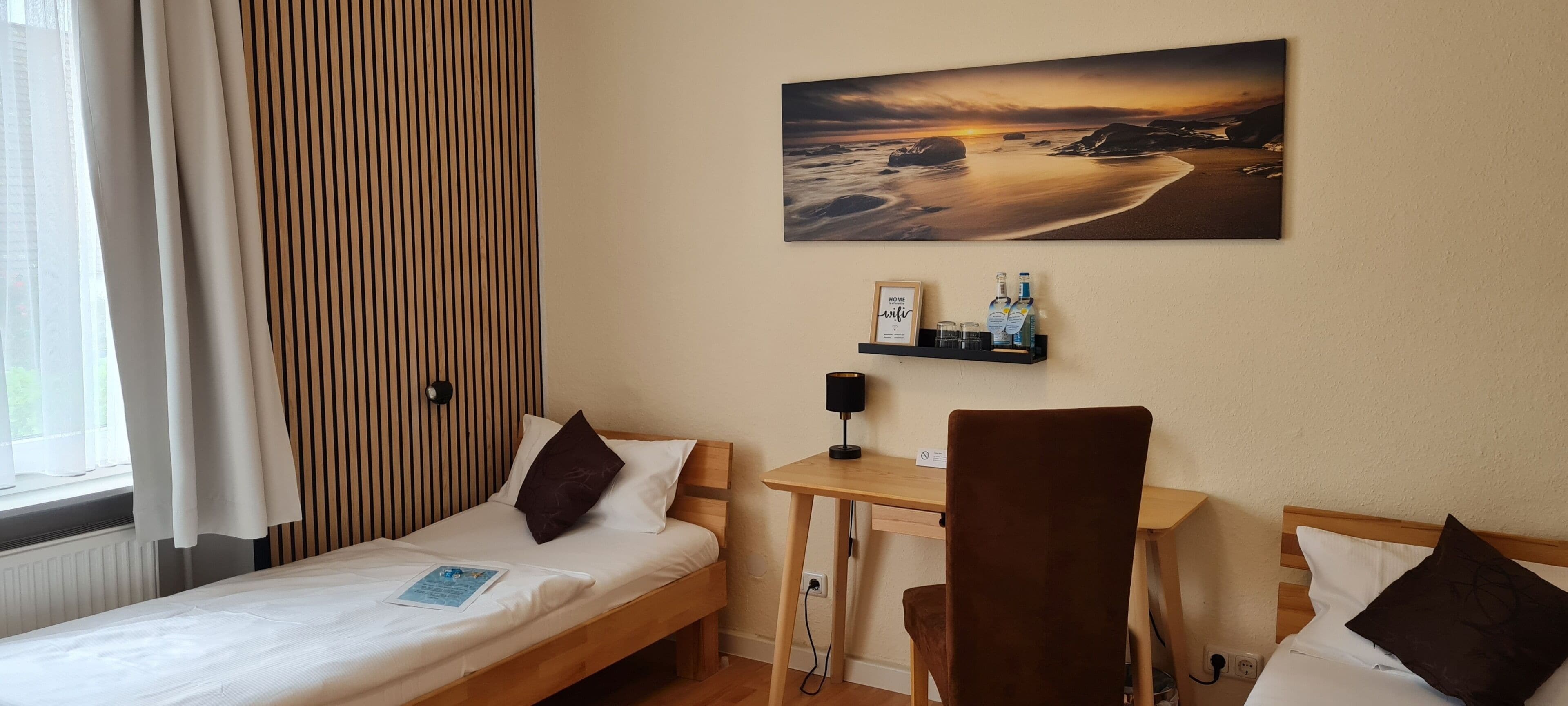 Una habitació d'hotel amb un llit, una taula de treball, una cadira, un quadre i una estant amb ampolles.