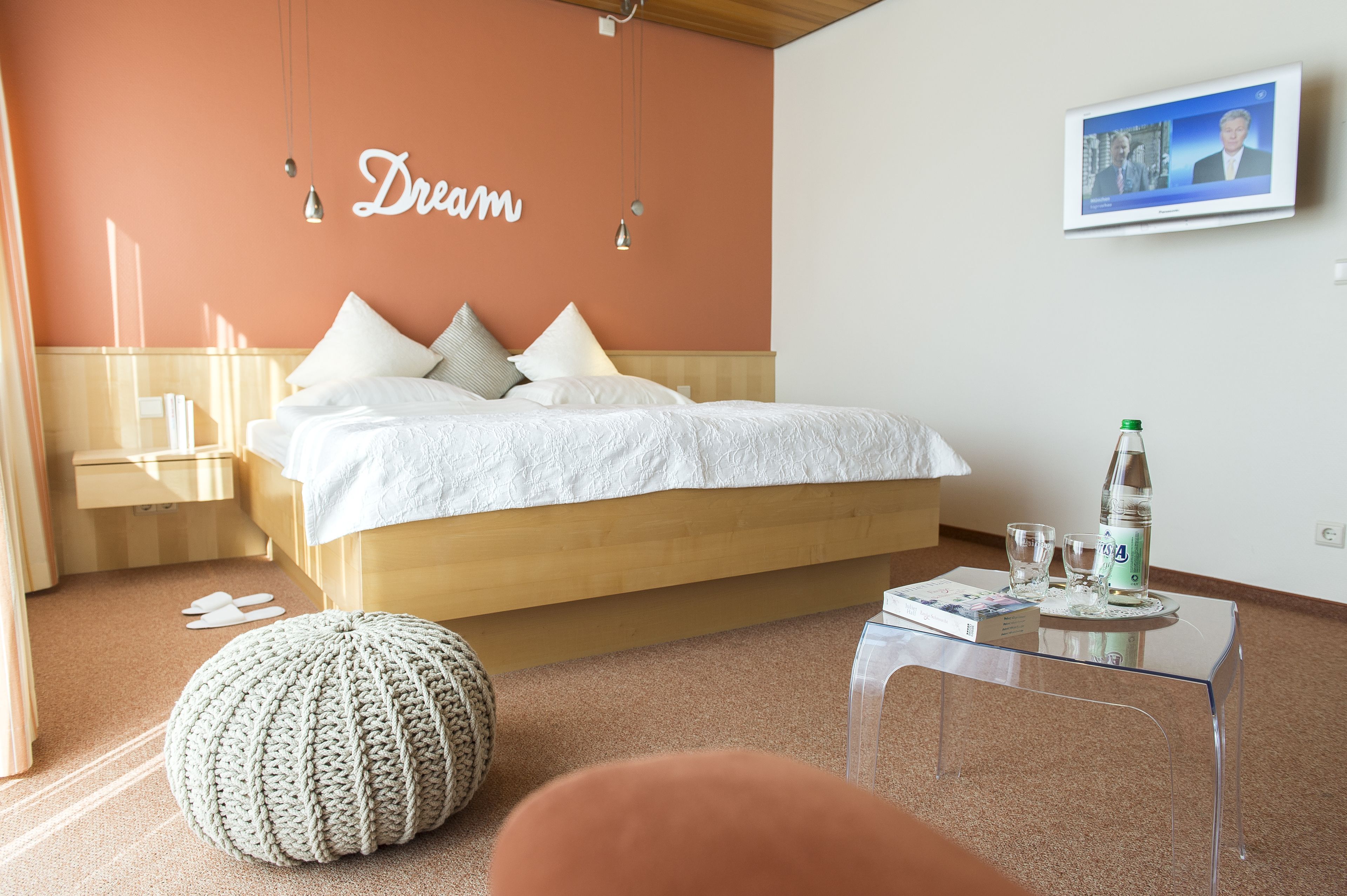 Ein Schlafzimmer mit einem Bett, einem Nachttisch, einem transparenten Glastisch, einem gestrickten Pouf und einer Wand mit der Aufschrift 'Dream'.
