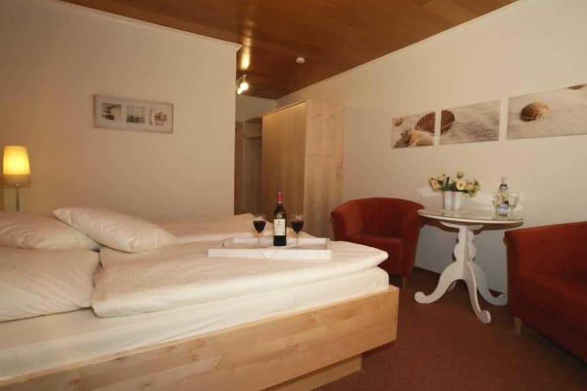 A bedroom with a wooden bed, a small table with a bottle and glasses, a red chair, and a wall with framed pictures.